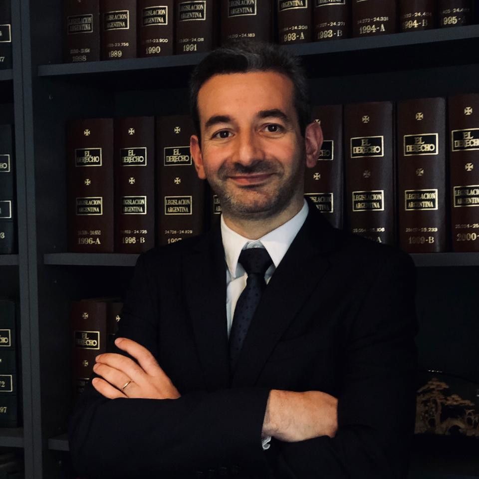 Néstor Javier Olijavetzky, de traje oscuro y brazos cruzados, ante la biblioteca jurídica del estudio